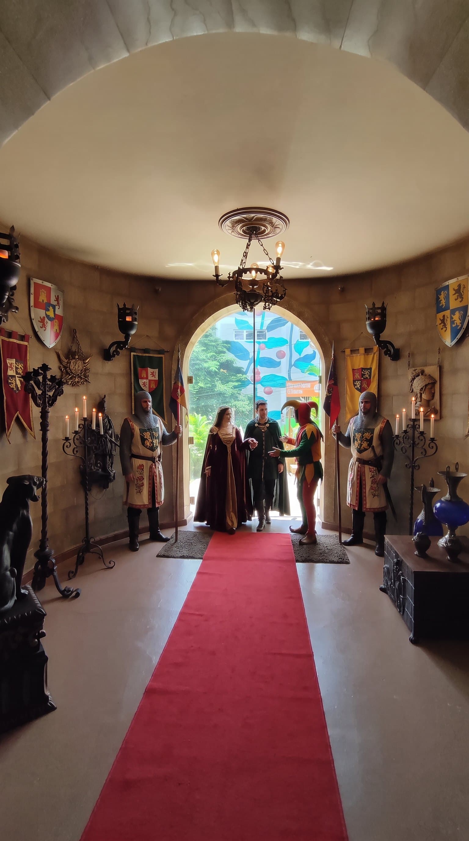Hall de entrada do castelo, com tapete vermelho, brasões nas paredes e candelabros de ferro